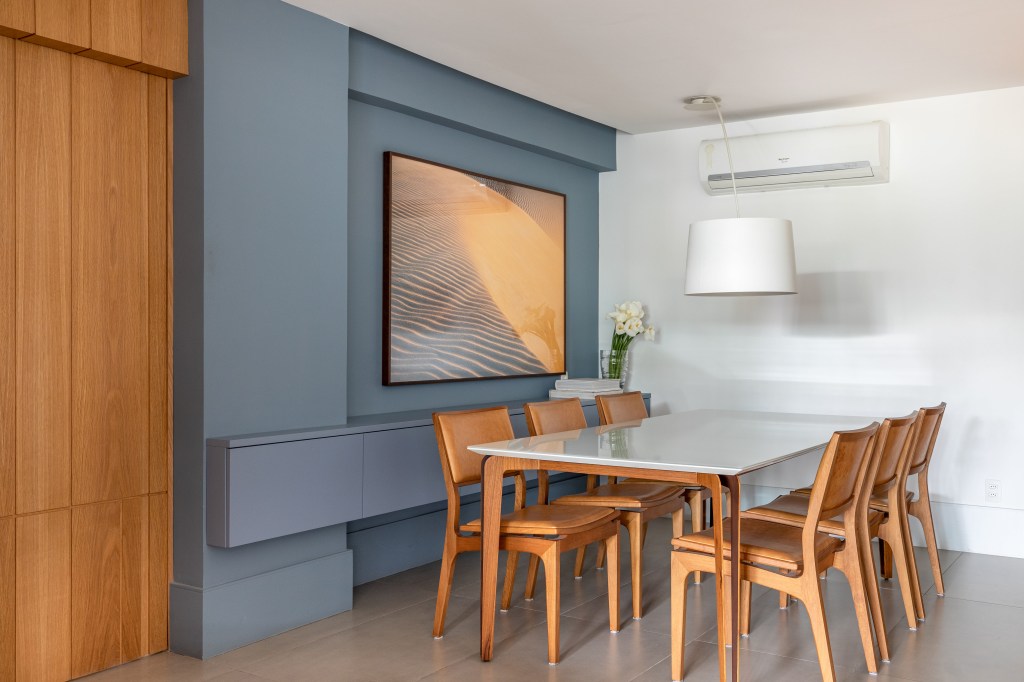 Sala de apê tem janela do piso ao teto para o terraço com jardineira. Projeto de Ao Quadrado Arquitetura. Na foto, sala de jantar, parede azul com quadro, mesa branca, cadeiras de madeira.