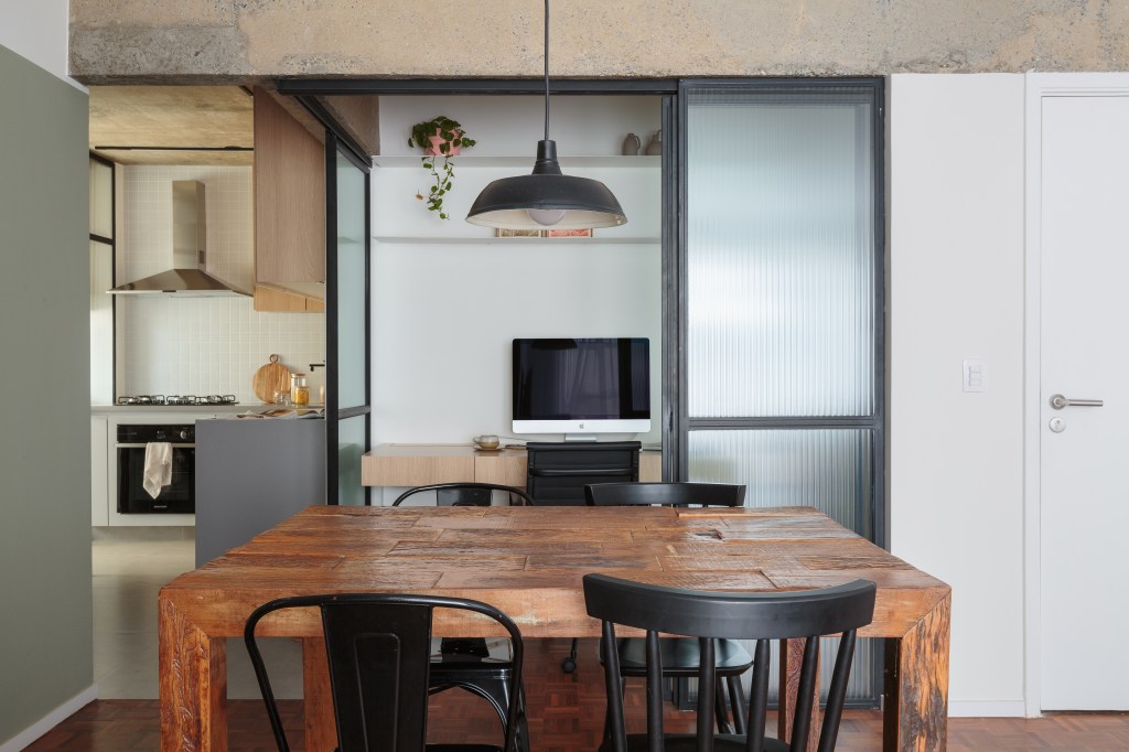 Apê de 70 m² ganha toque industrial com viga aparente e cama em metal. Projeto de Estúdio Maré. Na foto, sala de jantar com mesa de madeira, cadeiras pretas, home office com porta de vidro canelada, cozinha ao fundo.