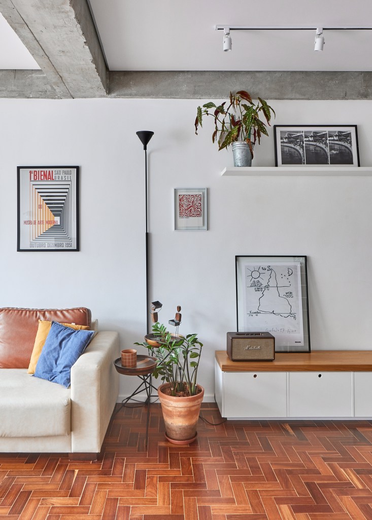 Vigas de concreto aparente emolduram área social de apê de 98 m². Projeto de Hugo Rapizo. Na foto, sala de estar, sofá branco, prateleira, rack baixo.