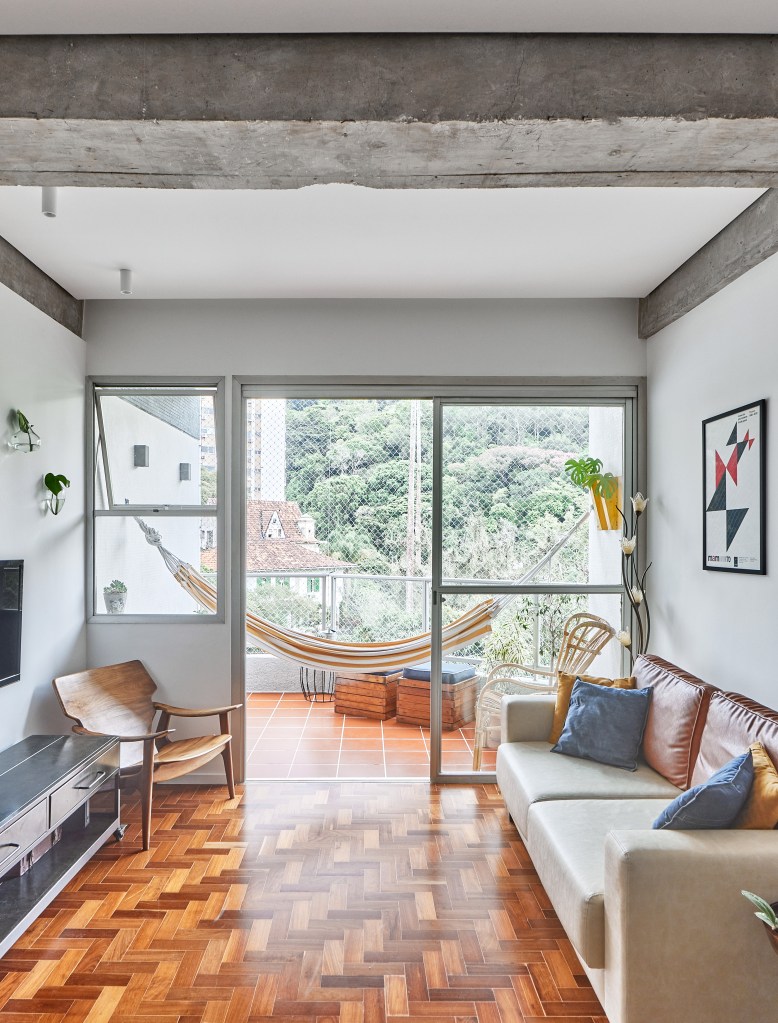 Vigas de concreto aparente emolduram área social de apê de 98 m². Projeto de Hugo Rapizo. Na foto, sala de estar, piso de taco, varanda com rede.